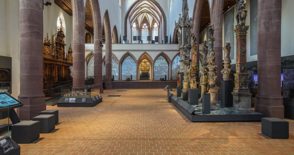 Blick vom Eingang in den Kircheninnenraum, Pfeiler aus rotem Sandstein unterteilen den Raum. Links vorne ein Computerbildschirm mit Sitzgelegenheiten davor, rechts in der Saalmitte sind auf einem Podest Beispiele von Brunnenstöcken aufgestellt.