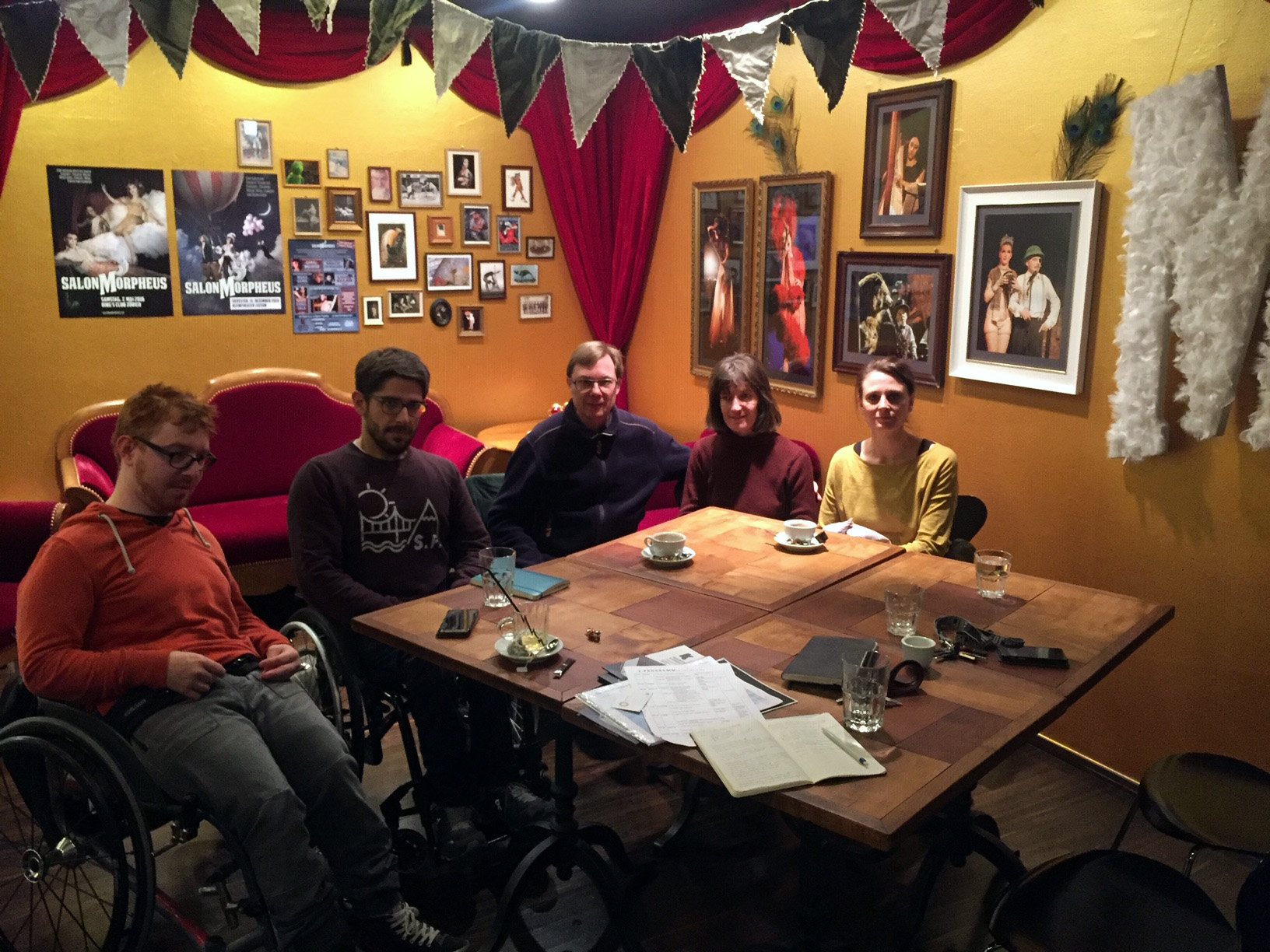 Fünf Personen sitzen um einen Tisch. Hinter ihnen ein Sofa und zwei Sessel, ein geraffter Theatervorhang und Volants an den Wänden, alles purpurrot und samtig. Theaterplakate und gerahmte Fotos von Bühnenauftritten. Fünf Personen sitzen um einen Tisch. Hinter ihnen ein Sofa und zwei Sessel, ein geraffter Theatervorhang und Volants an den Wänden, alles purpurrot und samtig. Theaterplakate und gerahmte Fotos von Bühnenauftritten.