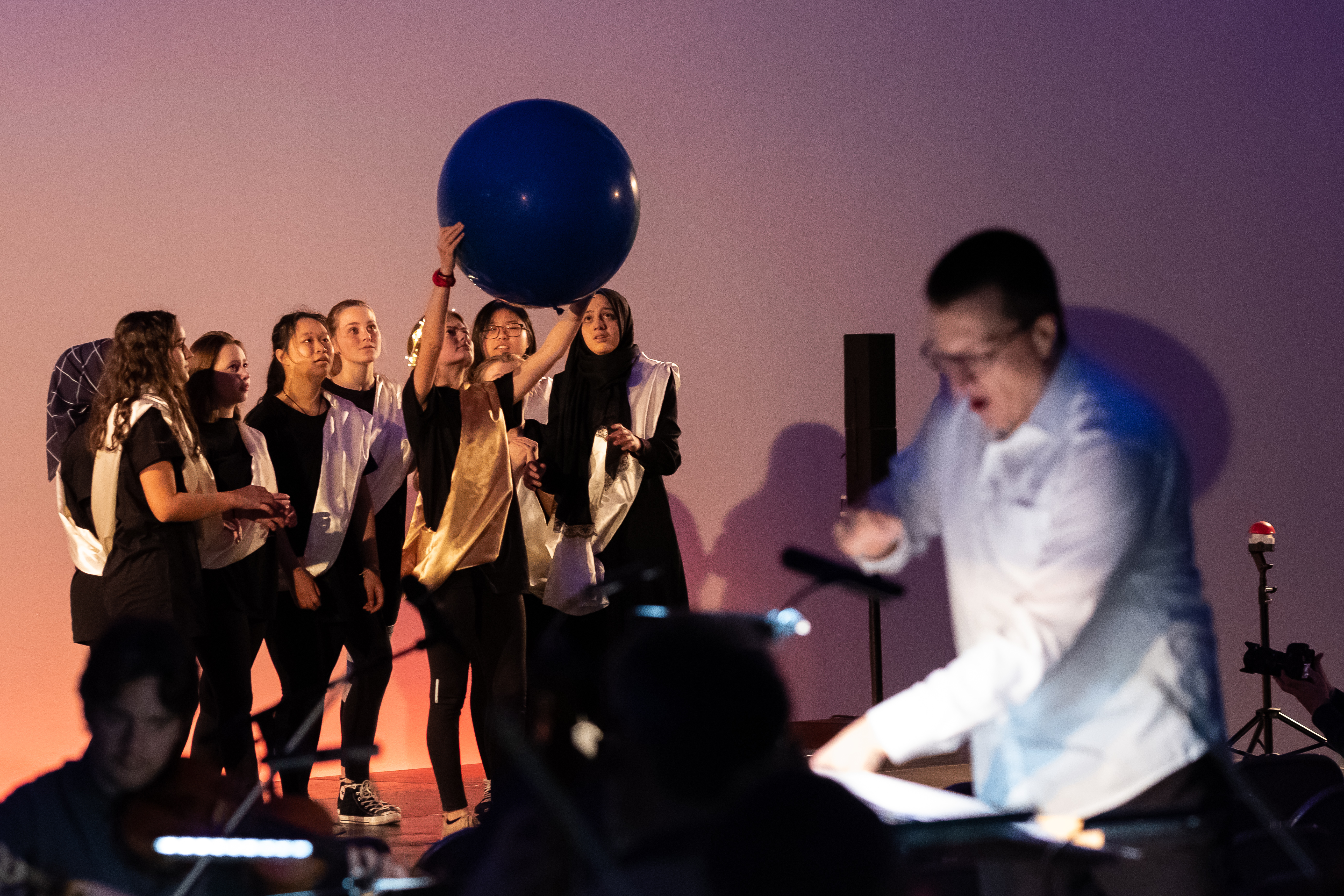Im Fokus eine Gruppe Jugendlicher auf einer Bühne. Schwarzgekleidet, mit einer hellen Schärpe, schauen sie zu einem grossen blauen Luftballon, den eine von ihnen hochhält. Im unscharfen Vordergrund ein Dirigent und ein Geiger.
