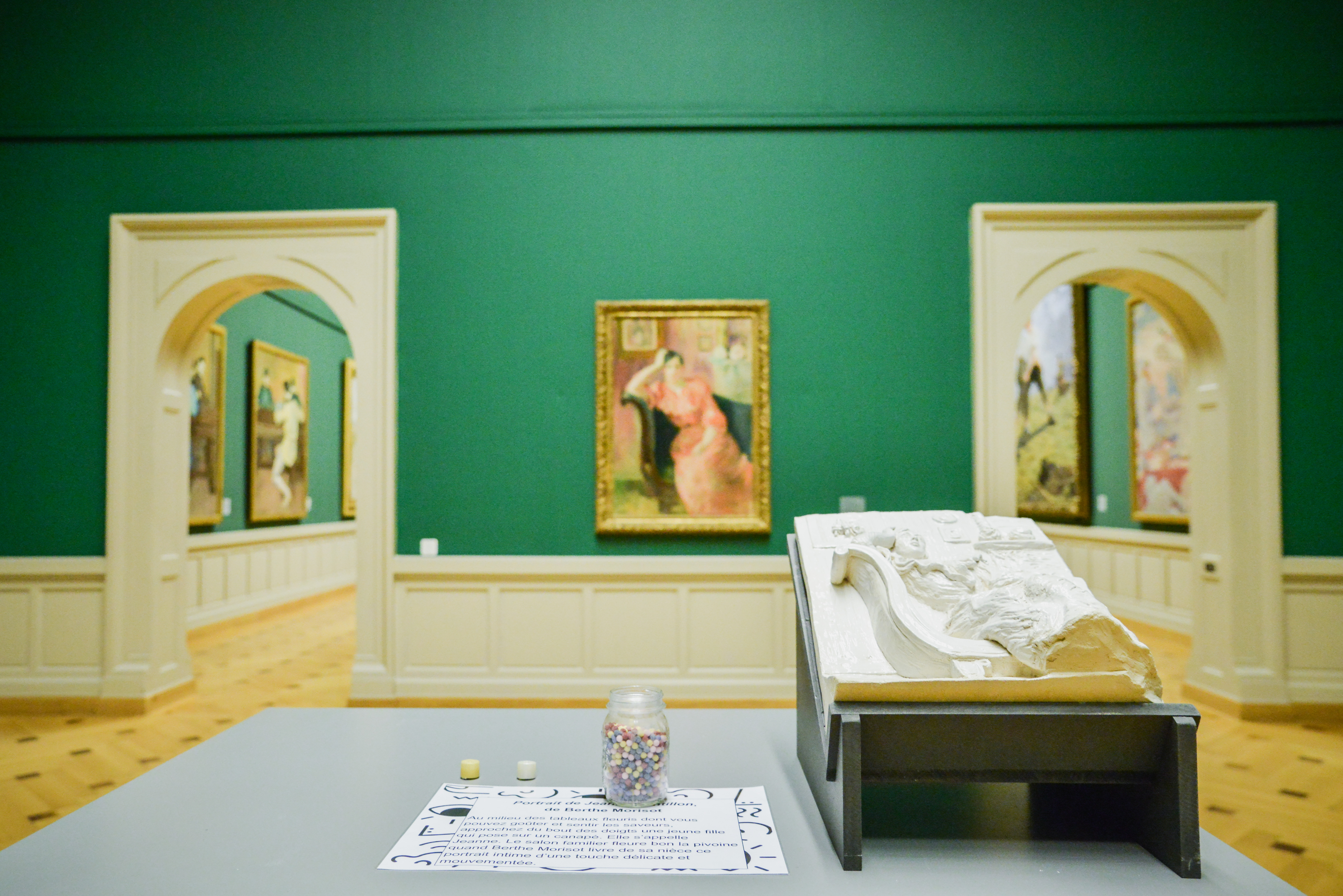 Sur une table, le bas-relief d’une femme sur un canapé. À côté, une bonbonnière remplie de billes de couleurs et une feuille avec du texte. Sur le mur d’en face, le portait de la femme. Par deux ouvertures, on voit des tableaux dans la salle suivante. Sur une table, le bas-relief d’une femme sur un canapé. À côté, une bonbonnière remplie de billes de couleurs et une feuille avec du texte. Sur le mur d’en face, le portait de la femme. Par deux ouvertures, on voit des tableaux dans la salle suivante.