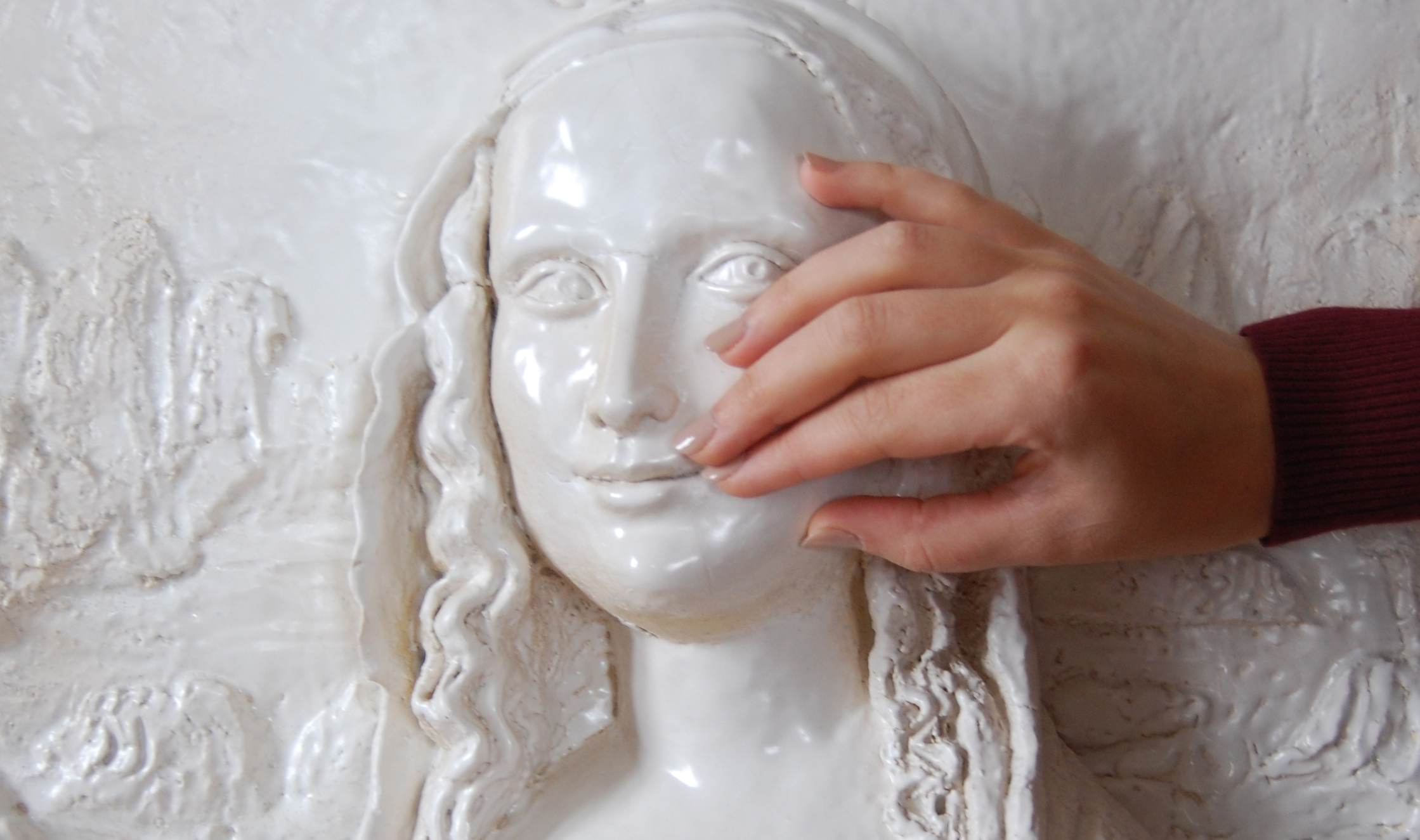 Bas-relief blanc brillant du visage et du cou d’une jeune femme aux traits réguliers, le regard tourné vers la droite, avec un léger sourire et de long cheveux ondulés. Une main touche la joue du bas-relief. Bas-relief blanc brillant du visage et du cou d’une jeune femme aux traits réguliers, le regard tourné vers la droite, avec un léger sourire et de long cheveux ondulés. Une main touche la joue du bas-relief.