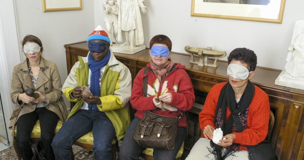 Vier Personen mit verbundenen Augen ertasten je ein kleines Objekt, das sie in den Händen halten. Sie sitzen nebeneinander auf brokatbezogenen, historischen Stühlen, auf dem Möbel mit Intarsien hinter ihnen stehen figürliche Marmorstatuetten. Vier Personen mit verbundenen Augen ertasten je ein kleines Objekt, das sie in den Händen halten. Sie sitzen nebeneinander auf brokatbezogenen, historischen Stühlen, auf dem Möbel mit Intarsien hinter ihnen stehen figürliche Marmorstatuetten.