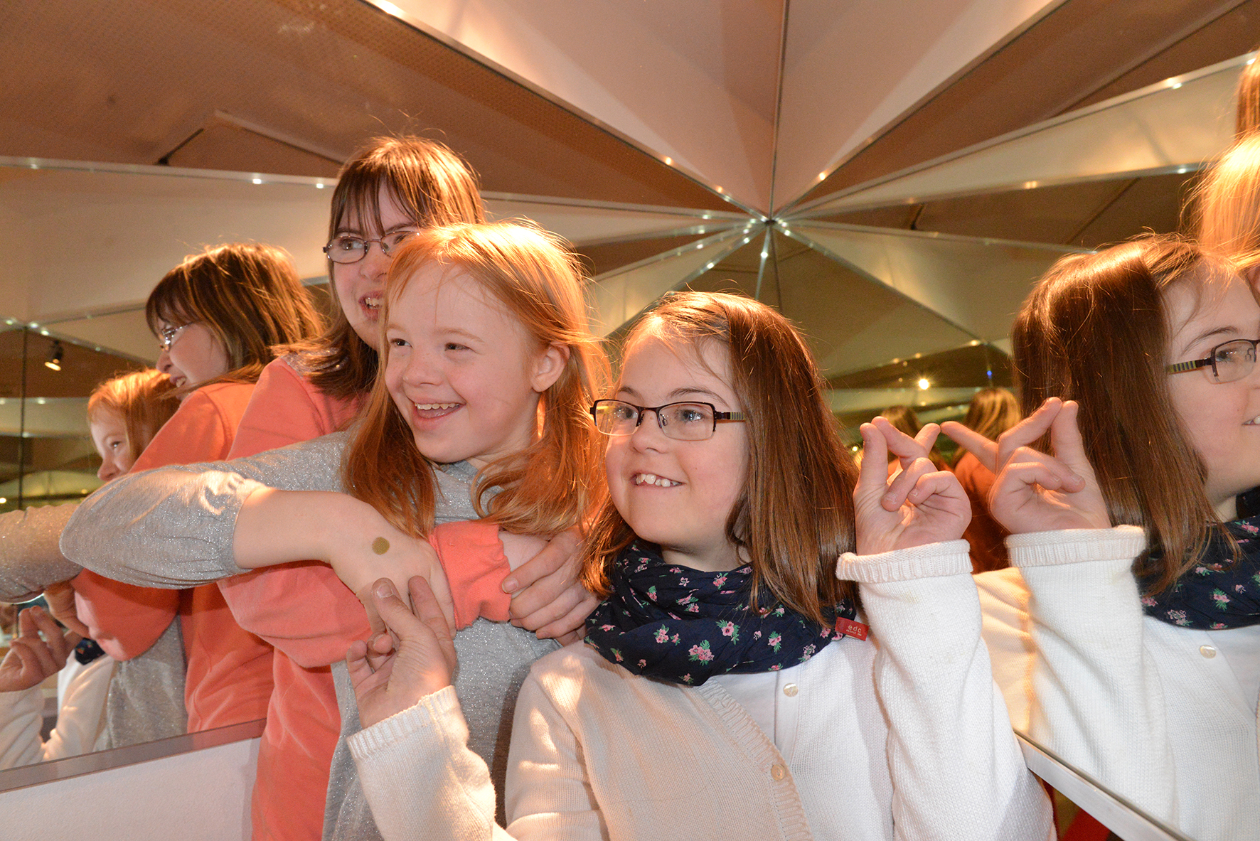 Drei Mädchen im Primarschulalter stehen dicht hintereinander in der Ecke eines Spiegelkabinetts, das sie vielfach widerspiegelt. Sie blicken lächelnd nach links. Das hinterste Mädchen umarmt das mittlere, das vordere zeigt auf die beiden dahinter.