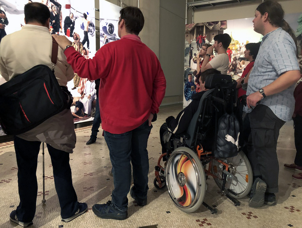 Des jeunes gens contemplent une installation photo de plusieurs mètres carré. Sur les photos, des adultes et des enfants de différents endroits du monde, et un avion.