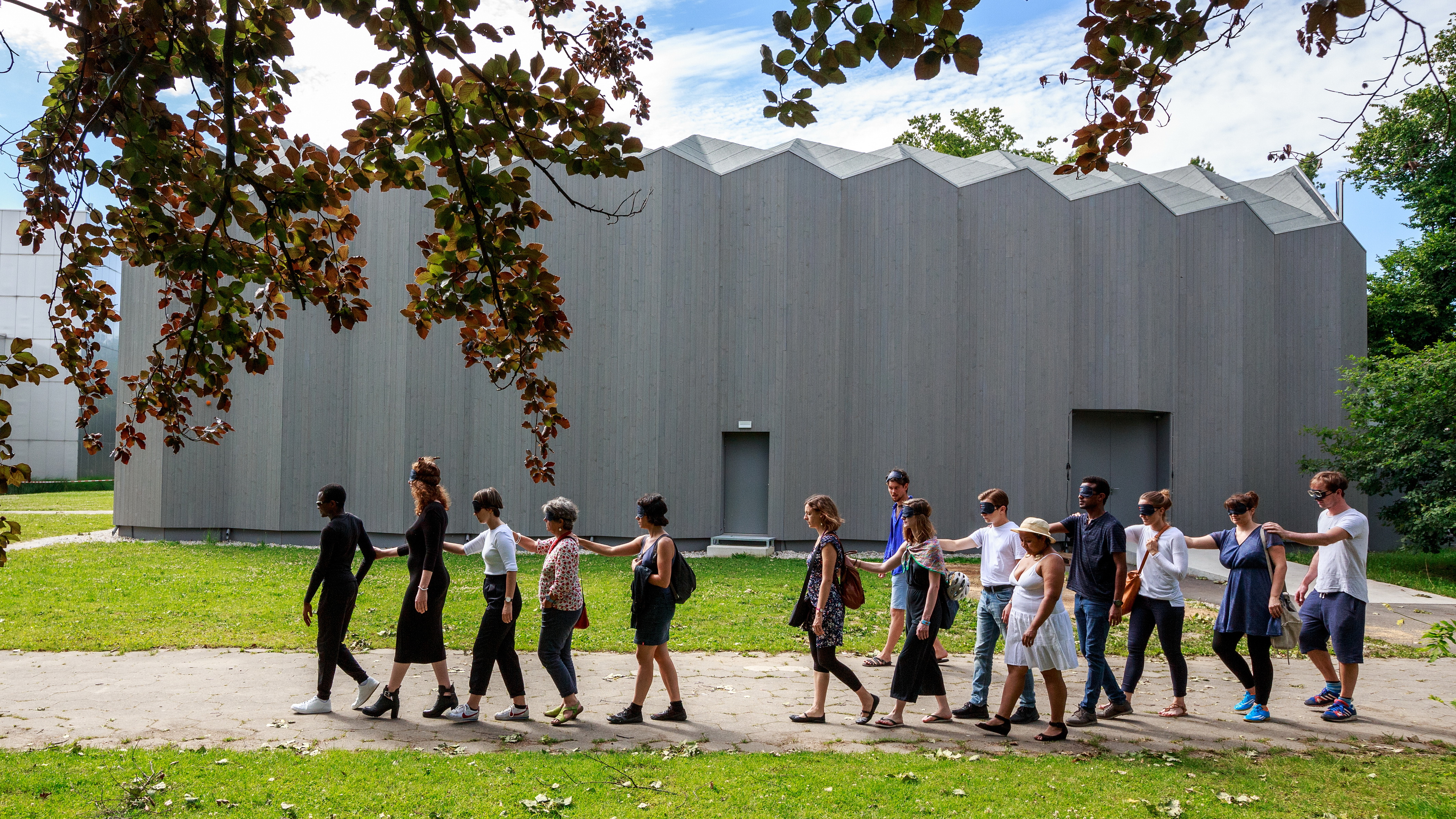 Une douzaine de personnes assez jeunes marchent en file indienne en se tenant par l’épaule. Presque toutes ont les yeux bandés. Derrière, un bâtiment gris sans fenêtre. Le chemin en terre battue sépare des pelouses. Au premier plan, des branches d’arbre. Une douzaine de personnes assez jeunes marchent en file indienne en se tenant par l’épaule. Presque toutes ont les yeux bandés. Derrière, un bâtiment gris sans fenêtre. Le chemin en terre battue sépare des pelouses. Au premier plan, des branches d’arbre.