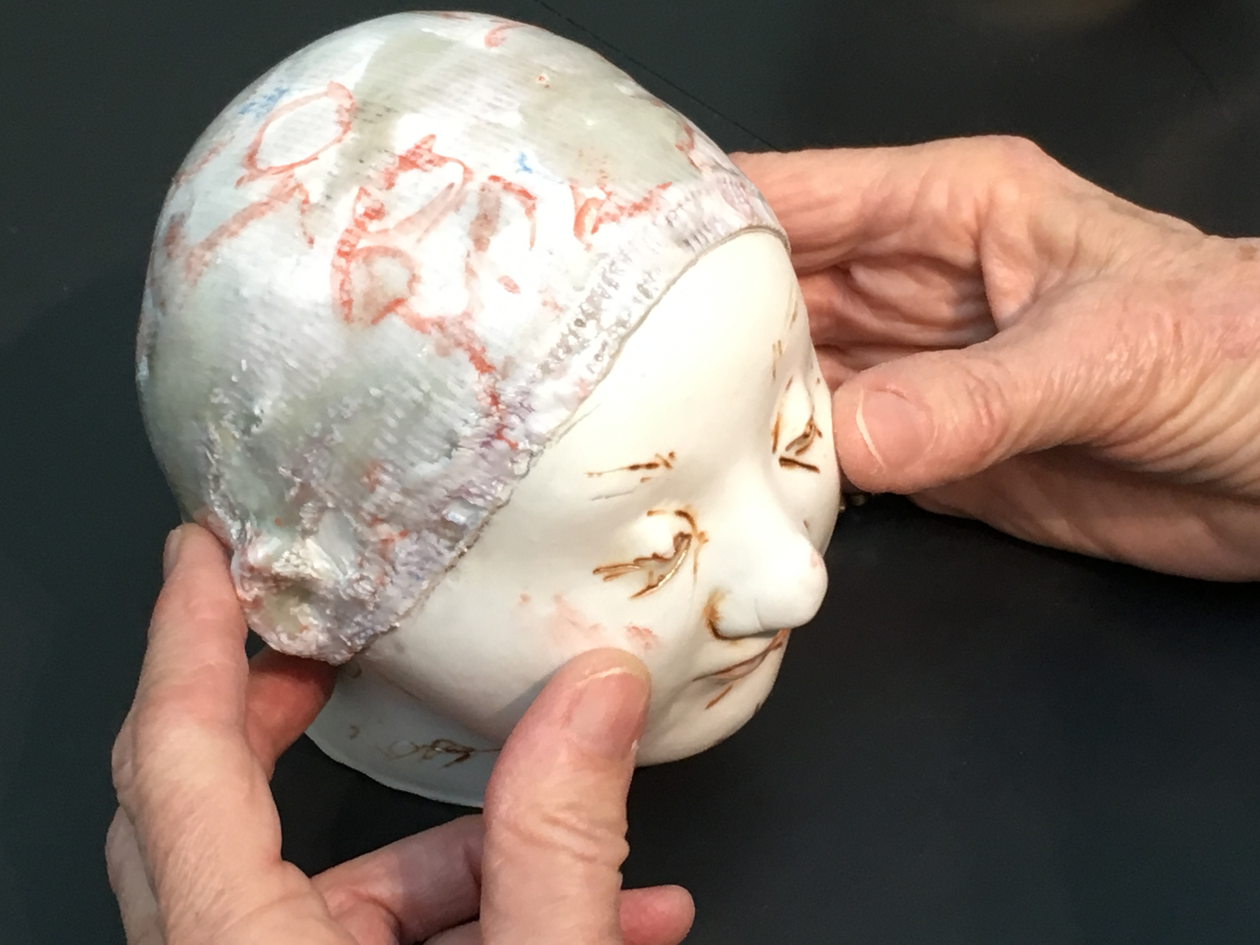 Des mains tiennent une tête sculptée de la taille d’un nourrisson. Des incisions beiges suggèrent les sourcils et soulignent les paupières baissées sur le visage blanc et lisse. La coiffe est rugueuse avec des taches grisâtres et des marques rouges. Des mains tiennent une tête sculptée de la taille d’un nourrisson. Des incisions beiges suggèrent les sourcils et soulignent les paupières baissées sur le visage blanc et lisse. La coiffe est rugueuse avec des taches grisâtres et des marques rouges.