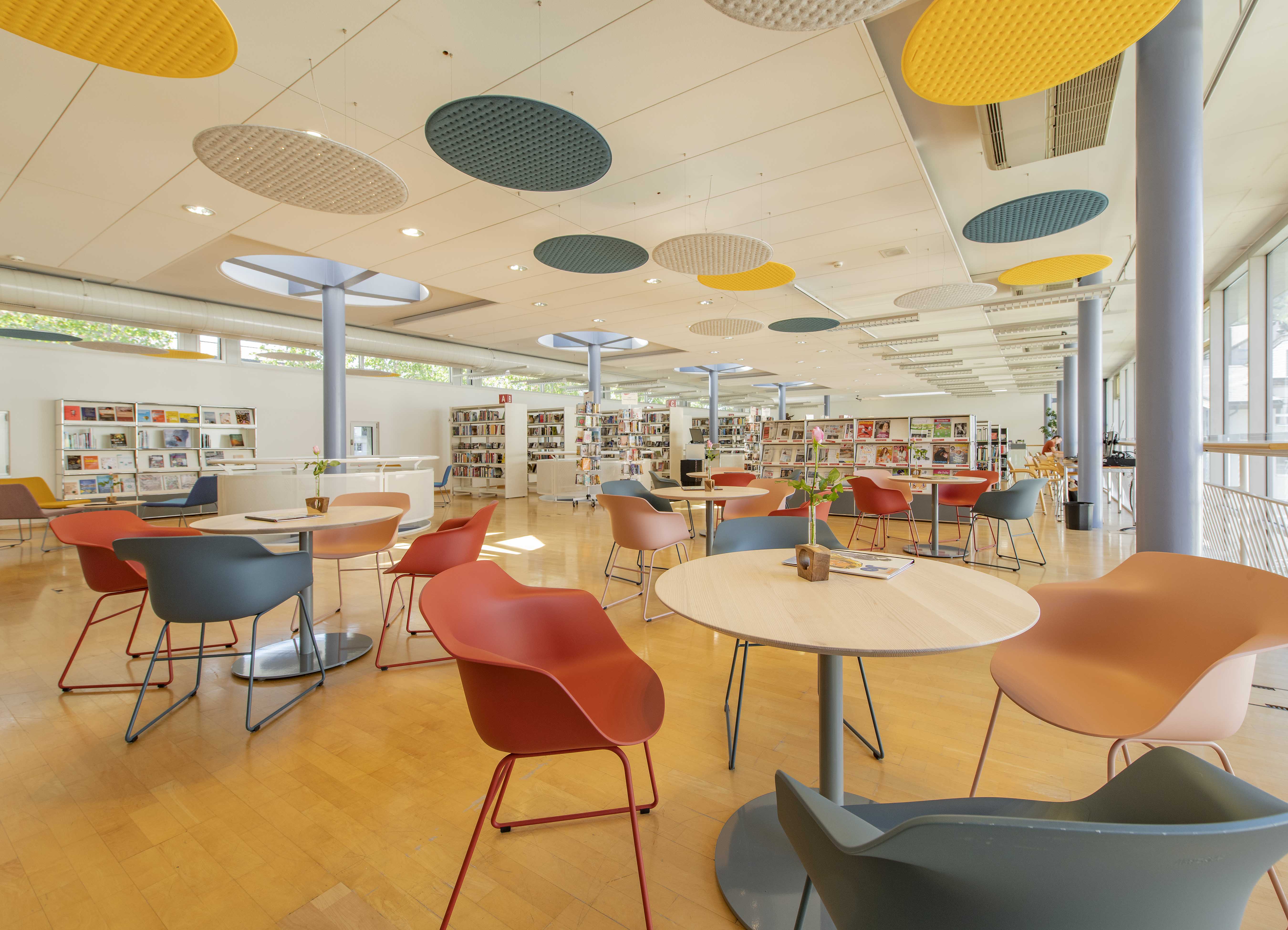 Eine Glasfassade und grosse, runde Oberlichter erhellen den ebenerdigen Raum. Vorne sind moderne Scha-lenstühle in Rot, Apricot und Petrol um Bistrotische drapiert, hinten stehen Bücherregale. An der Decke sind runde Schalldämmer in Gelb, Petrol, Weiss. Eine Glasfassade und grosse, runde Oberlichter erhellen den ebenerdigen Raum. Vorne sind moderne Scha-lenstühle in Rot, Apricot und Petrol um Bistrotische drapiert, hinten stehen Bücherregale. An der Decke sind runde Schalldämmer in Gelb, Petrol, Weiss.