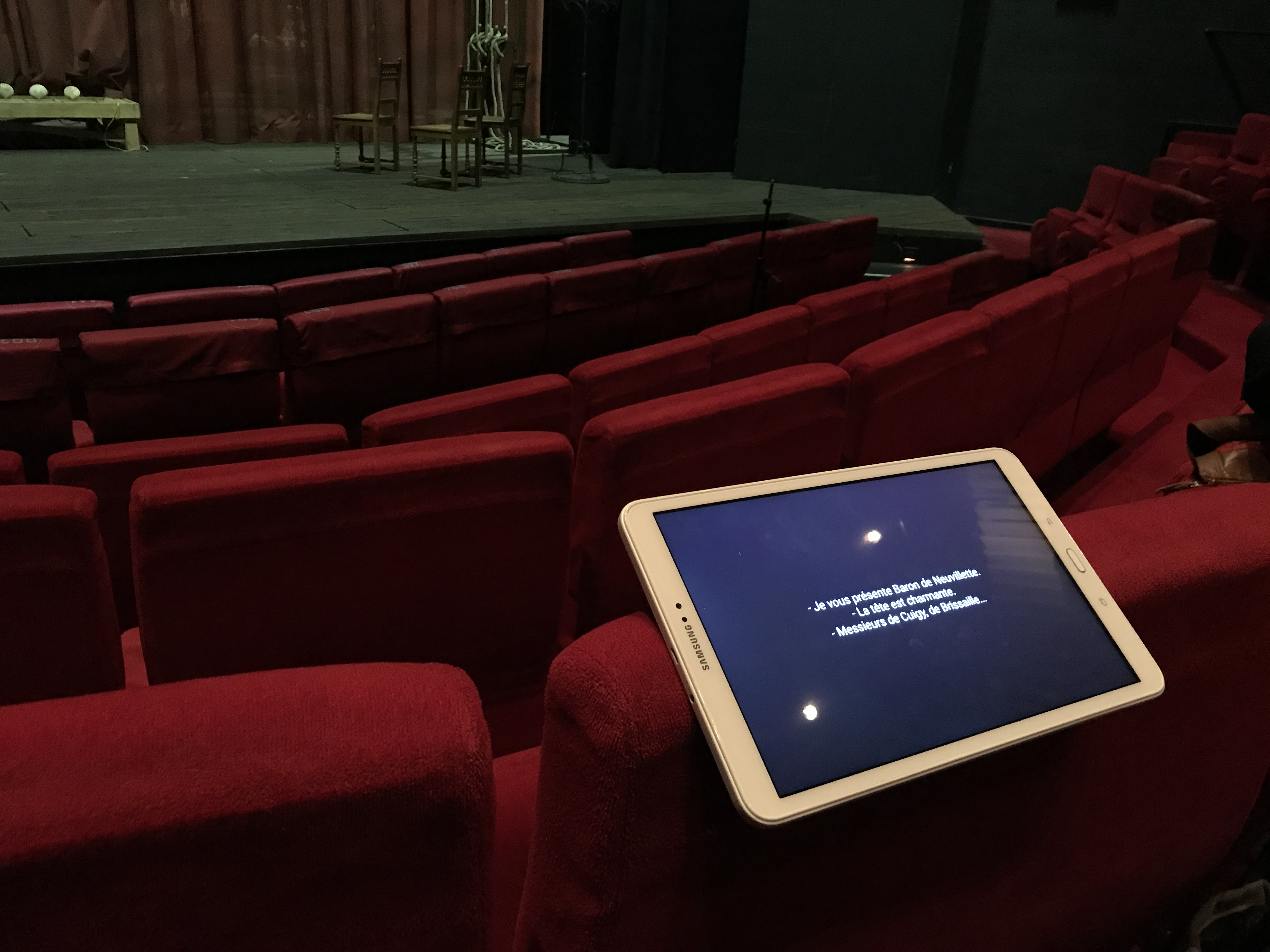 Une tablette posée en équilibre sur le dossier d’un siège en velours rouge dans une salle de théâtre vide. Au se-cond plan, la scène. La tablette est allumée, trois courtes phrases s’affichent sur l’écran. Une tablette posée en équilibre sur le dossier d’un siège en velours rouge dans une salle de théâtre vide. Au se-cond plan, la scène. La tablette est allumée, trois courtes phrases s’affichent sur l’écran.