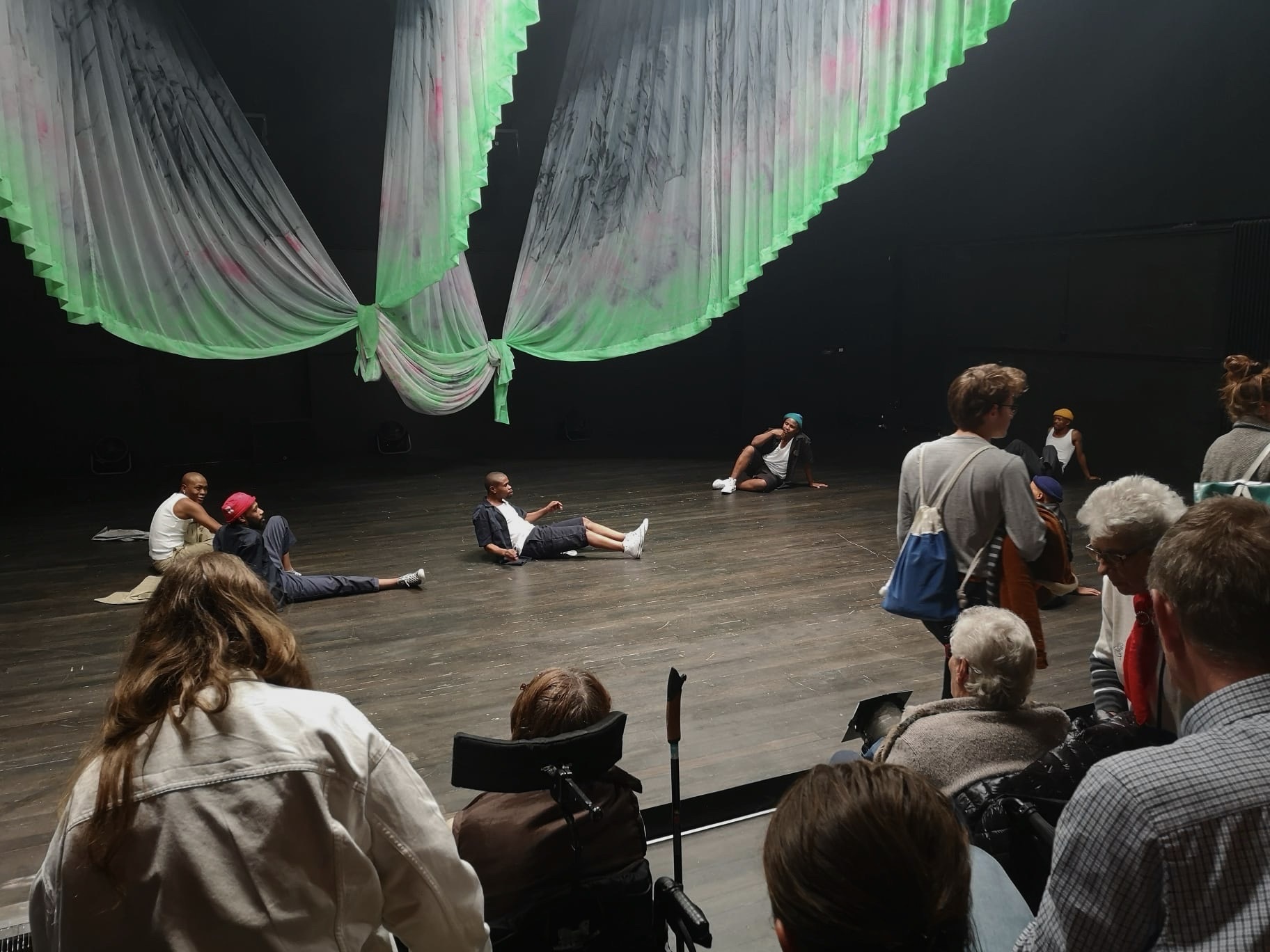 Un public d’âge varié, assis, debout ou en train de se déplacer dans les gradins. Sur la scène, six jeunes hommes, confortablement assis au sol, se regardent. Des bandes de tissu pendent du plafond comme des ailes, l’éclairage de la scène est uniforme. Un public d’âge varié, assis, debout ou en train de se déplacer dans les gradins. Sur la scène, six jeunes hommes, confortablement assis au sol, se regardent. Des bandes de tissu pendent du plafond comme des ailes, l’éclairage de la scène est uniforme.