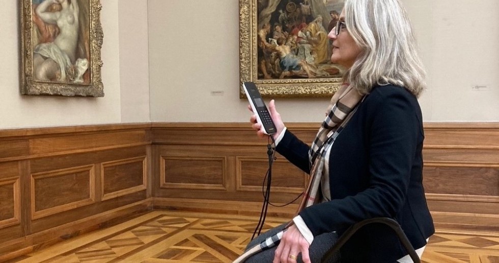 Die Ecke eines Raumes mit zwei goldgerahmten Gemälden, Holzparkett mit Intarsien und hüfthoher Leiste. In der Saalmitte sitzt eine Frau mit schulterlangen grauen Haaren auf einem Klapphocker, einen Audioguide in der Hand und blickt zur Wand gegenüber.