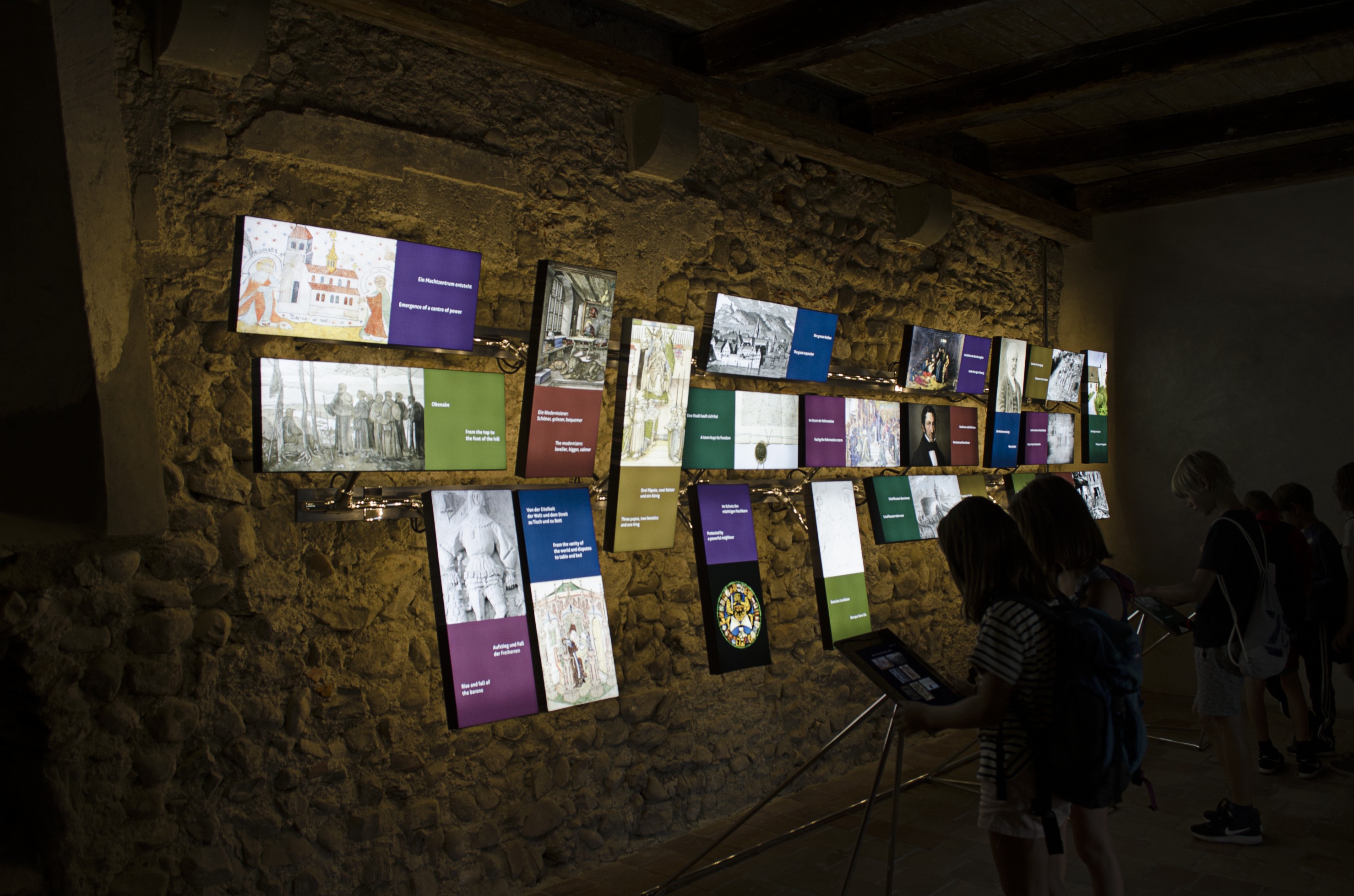 Dunkler, höhlenartiger Ausstellungsraum, eine dunkle, gewölbte Steinwand. Daran sind beleuchtete kleine Bilder im Hoch- und Querformat angebracht. Ausstellungsbesucher*innen studieren Anzeigen vor der Wand.
