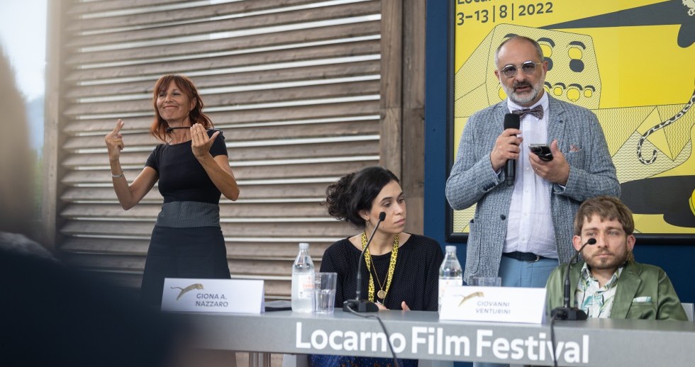 Zwei Personen sitzen an einem Tisch auf dem «Locarno Film Festival» prangt, Mikrofone und Namensschildern vor ihnen. Zwischen ihnen steht ein Mann mit Mikrofon in der Hand. Dahinter hängt das Festivalplakat. Links gebärdet eine schwarzgekleidete Frau. Zwei Personen sitzen an einem Tisch auf dem «Locarno Film Festival» prangt, Mikrofone und Namensschildern vor ihnen. Zwischen ihnen steht ein Mann mit Mikrofon in der Hand. Dahinter hängt das Festivalplakat. Links gebärdet eine schwarzgekleidete Frau.