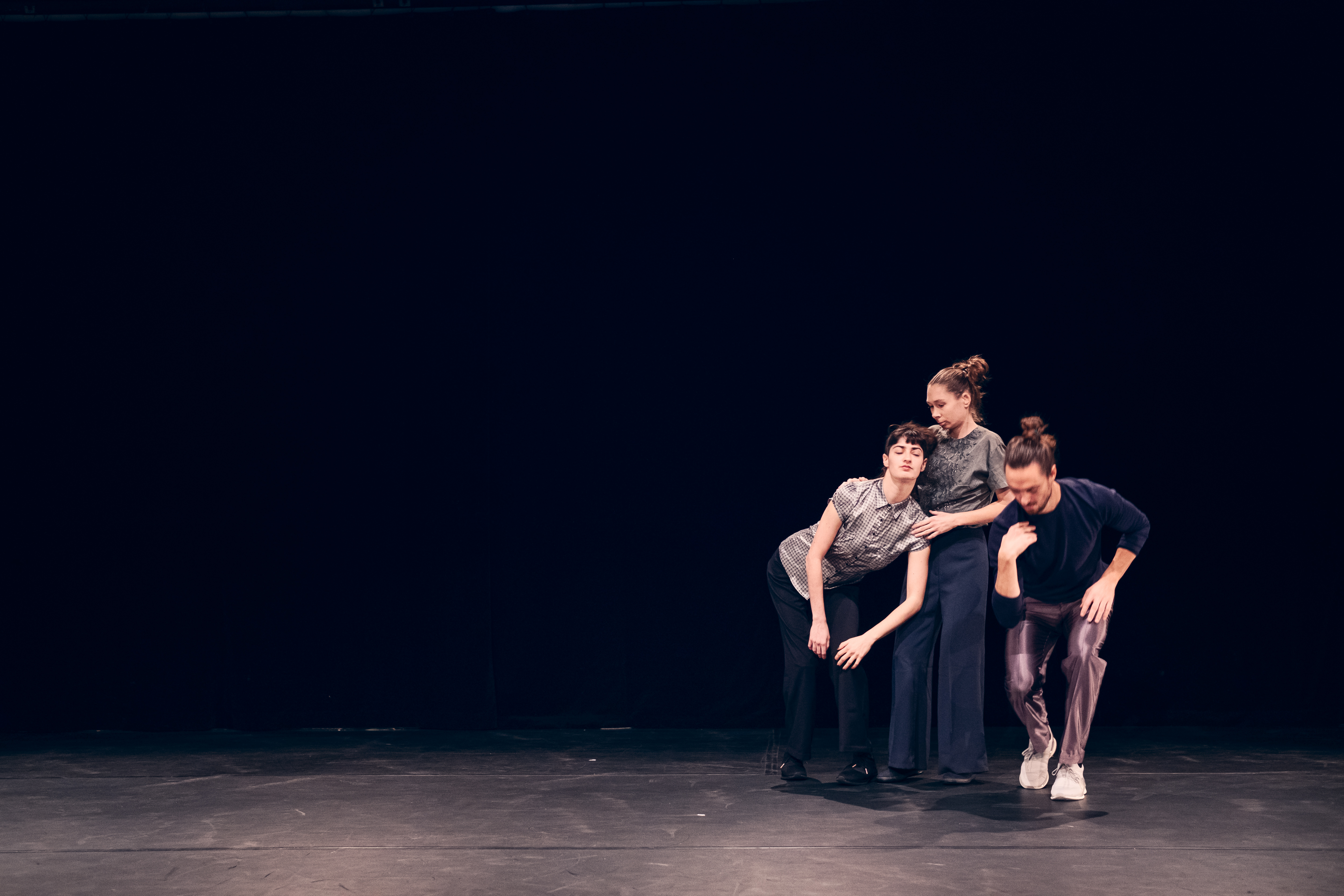 Drei Performer*innen nebeneinander auf der Bühne vor schwarzem Hintergrund. Eine lehnt sich mit geschlossen Augen an die andere. Diese steht aufrecht, ihr zugewandt, die Hände auf ihren Schultern. Die dritte beugt Oberkörper und Knie, Blick nach unten.