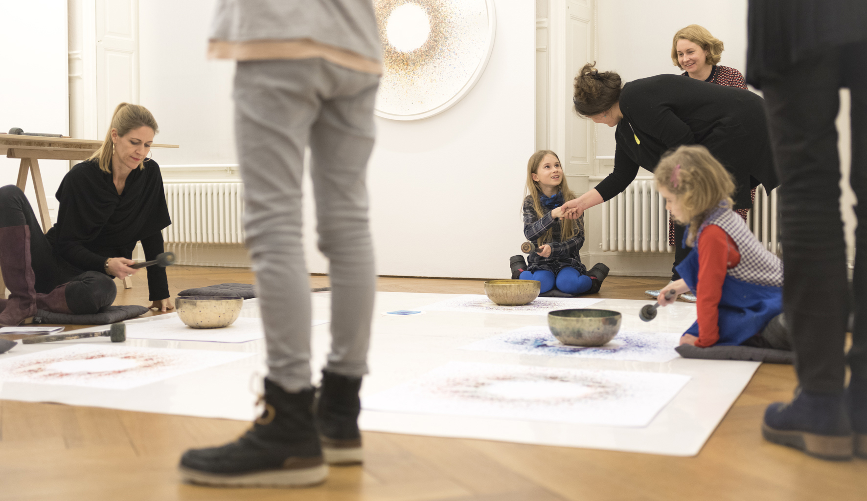 Eine Erwachsene und zwei Kinder auf dem Boden an den Rändern einer Abdeckfolie, einen Schlegel in der Hand, eine Klangschüssel vor sich auf einem Blatt Papier. Um die Schüssel sind kreisförmig Farbtupfer gestreut. Ein ähnliches Sujet hängt an der Wand.
