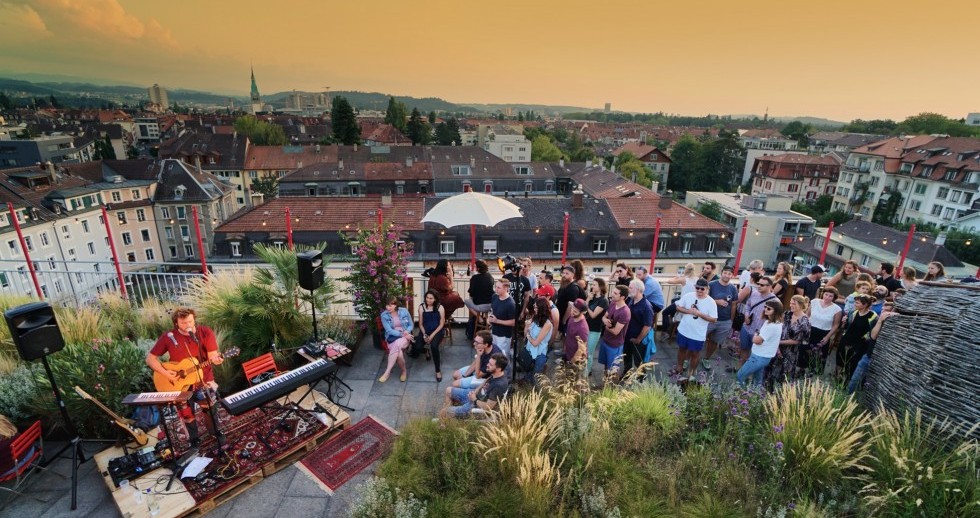Auf einer begrünten Dachterrasse verfolgen über vierzig Menschen unterschiedlichen Alters ein Konzert eines Musikers mit Gitarre. Er steht auf einer Bühne aus Bahnpaletten zwischen zwei Keybords und einer Elektrogitarre sowie Mischpult und zwei Boxen.