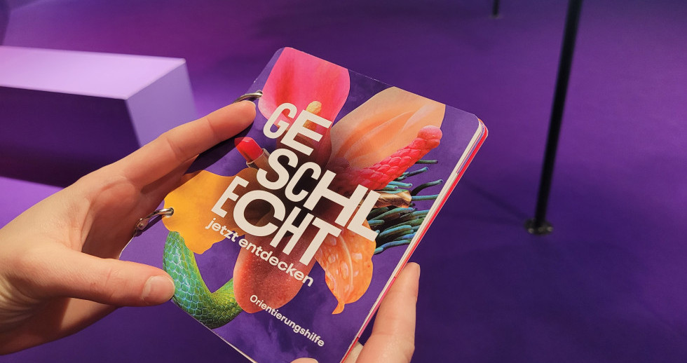 In der Bildmitte halten zwei Hände ein Büchlein mit dem Blüten-Sujet der Ausstellung und der Aufschrift «GESCHLECHT – jetzt entdecken. Orientierungshilfe». Die Blüte ist rosa-orange, der Hintergrund violett, wie der Ausstellungsraum im Hintergrund.