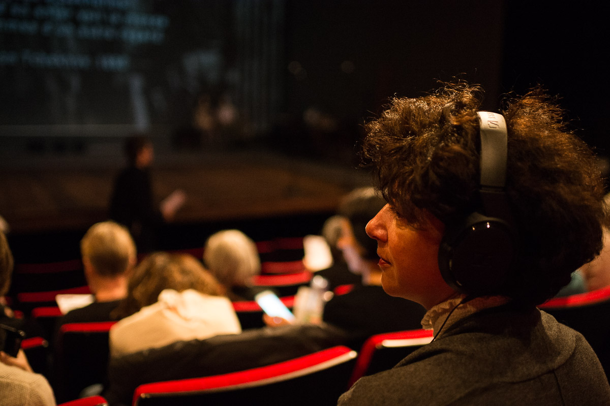 Au premier plan, une personne de dos, le visage de profil, avec un casque sur les oreilles. Elle est assise dans les gradins d’une salle de théâtre. La scène est peu éclairée et vide.