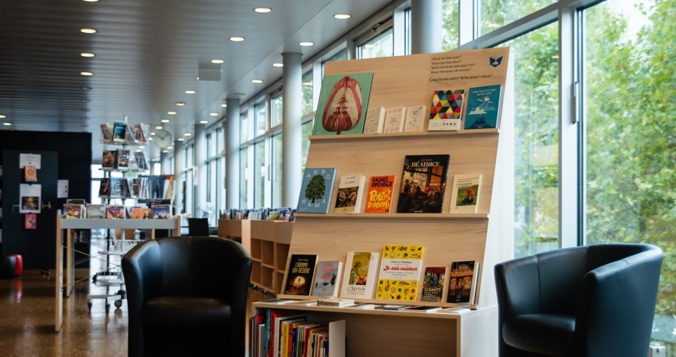 Adossé à une baie vitrée qui donne sur des arbres verts, un présentoir à livres en bois clair sur roulettes, incliné vers l'arrière et flanqué de deux fauteuils noirs, dans un espace au parquet clair. Les livres ont des couvertures colorées.