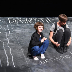 Une enfant et une femme sont assises sur une scène. Elle a la forme d’un tableau noir incliné vers l’avant. Il y a des dessins à la craie : un corps sans tête et les inscriptions « diagnostic » et « normale ». Au fond à droite il y a une pile de livres.
