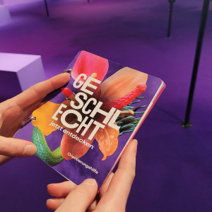 In der Bildmitte halten zwei Hände ein Büchlein mit dem Blüten-Sujet der Ausstellung und der Aufschrift «GESCHLECHT – jetzt entdecken. Orientierungshilfe». Die Blüte ist rosa-orange, der Hintergrund violett, wie der Ausstellungsraum im Hintergrund.