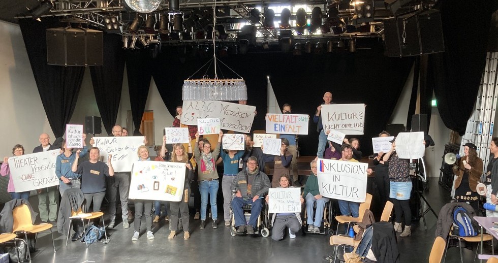 Jüngere und ältere Menschen mit und ohne Behinderungen schauen in die Kamera, lächeln und halten handgeschriebene Plakate hoch. Darauf steht etwa «Vielfalt statt Einfalt» oder «Kultur ist bunt». Sie sind auf einer Bühne in einem Theatersaal.