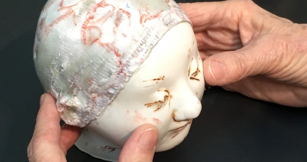 Hände halten einen modellierten Kopf in der Grösse eines Säuglings. Beige Einschnitte deuten Augenbrauen an und betonen die gesenkten Augenlider auf dem glatten weissen Gesicht. Die Kopfbedeckung ist rau mit gräulichen Flecken und roten Markierungen. Hände halten einen modellierten Kopf in der Grösse eines Säuglings. Beige Einschnitte deuten Augenbrauen an und betonen die gesenkten Augenlider auf dem glatten weissen Gesicht. Die Kopfbedeckung ist rau mit gräulichen Flecken und roten Markierungen.