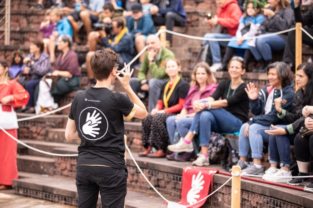 Ein Mann steht in einem römischen Theater. Ein Symbol mit zwei Händen ist auf dem Rücken seines T-Shirts aufgedruckt: das internationale Symbol für Gebärdensprache. Das T-Shirt macht ihn als Dolmetscher für Gebärdensprache erkennbar. Seine Hände bilden Ge