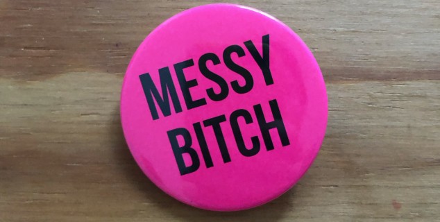 Das Bild zeigt einen pinken Button. Der Button ist rund und hat etwa einen Durchmesser von sieben Zentimetern. Er trägt eine schwarze Aufschrift, die sagt: Messy Bitch (deutsch: chaotische Schlampe).