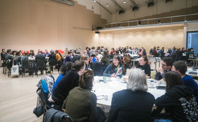 Ein grosser Saal mit hoher Decke. Im Raum verteilt sind mehrere runde Tische, an denen circa zehn bis zwölf Personen sitzen. Am Tisch im Vordergrund sind die Blicke auf eine Frau gerichtet, die am Sprechen ist und ein Buch in der Hand hält.