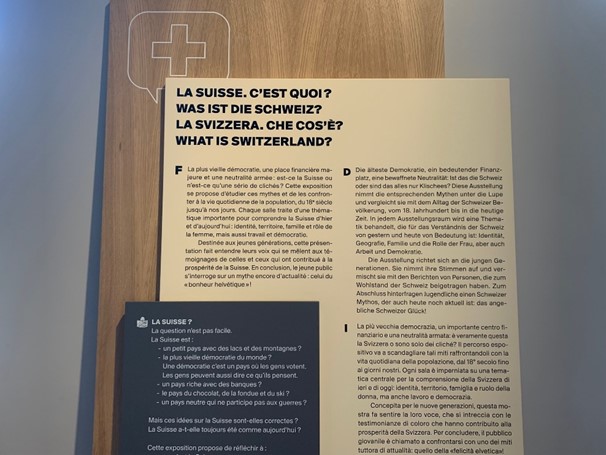 Un panneau avec trois blocs de textes noirs sur fond blanc et un texte blanc sur fond noir. Le titre « La Suisse. C’est quoi ? » est écrit en français, allemand, italien et anglais. En haut du panneau, le contour d’un écusson avec une croix. Un panneau avec trois blocs de textes noirs sur fond blanc et un texte blanc sur fond noir. Le titre « La Suisse. C’est quoi ? » est écrit en français, allemand, italien et anglais. En haut du panneau, le contour d’un écusson avec une croix.