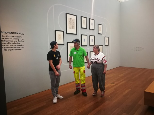 Drei Personen stehend in einem von Oberlicht erhelltem Saal. An der grauen Wand hinter ihnen sind ein Dutzend schwarzgerahmte Bleistiftskizzen gruppiert. Zwei Personen halten sich an der Hand und sind der dritten zugewandt, die zu ihnen spricht.