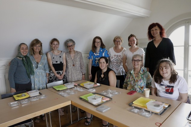 Dans un espace intérieur, 11 femmes debout ou assises à deux tables sur lesquelles sont posés des plateaux, des récipients avec du liquide ou une matière blanche, des éponges et des carrés de tissu épais et colorés. 