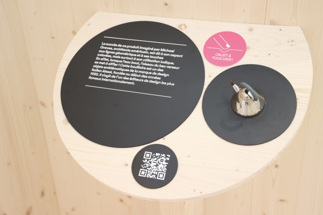 Une tablette en demi-cercle sur laquelle sont représentés quatre cercles. Le premier comporte un bref texte, le deuxième un QR code, le troisième une bouilloire miniature et le dernier la mention « Objet à toucher ! » avec un pictogramme de doigt. 